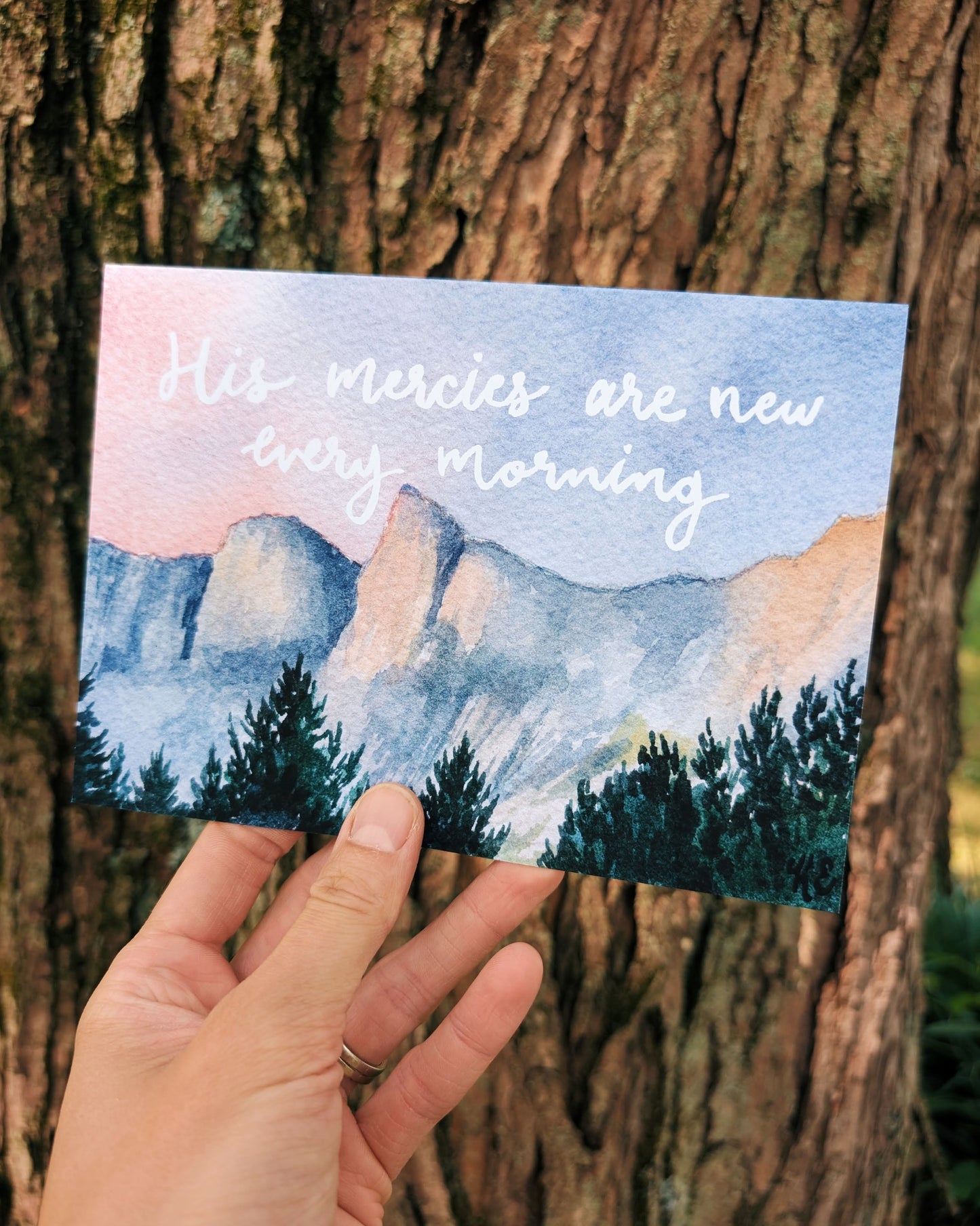 Hand holding a card with a mountain landscape and 'His mercies are new every morning' text against a mountain background.