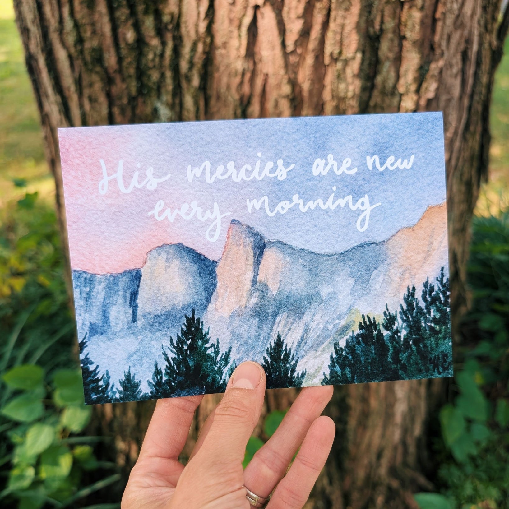 Hand holding a postcard with a mountain landscape and 'His mercies are new every morning' text against a mountain background.