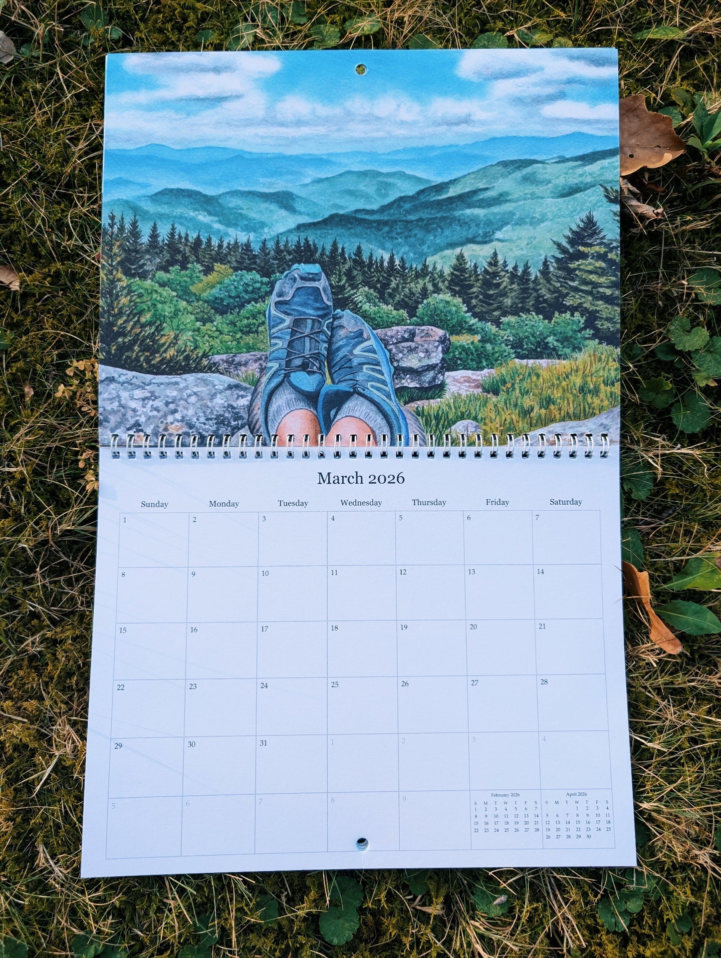 Calendar with a scenic illustration of a person in hiking boots overlooking a mountain landscape, placed on grass.