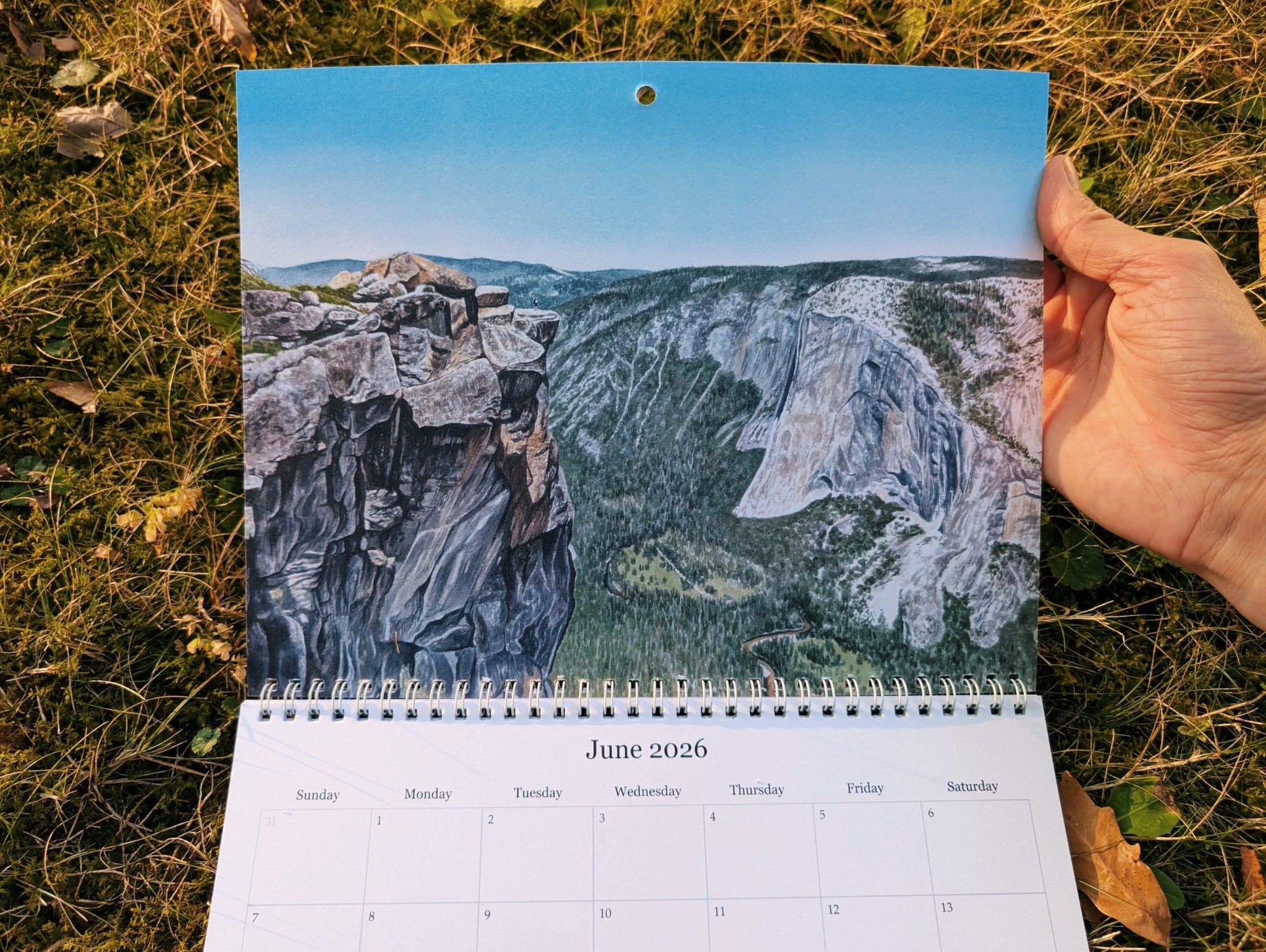 Calendar with a watercolor painting of Yosemite national park held by a hand on grass