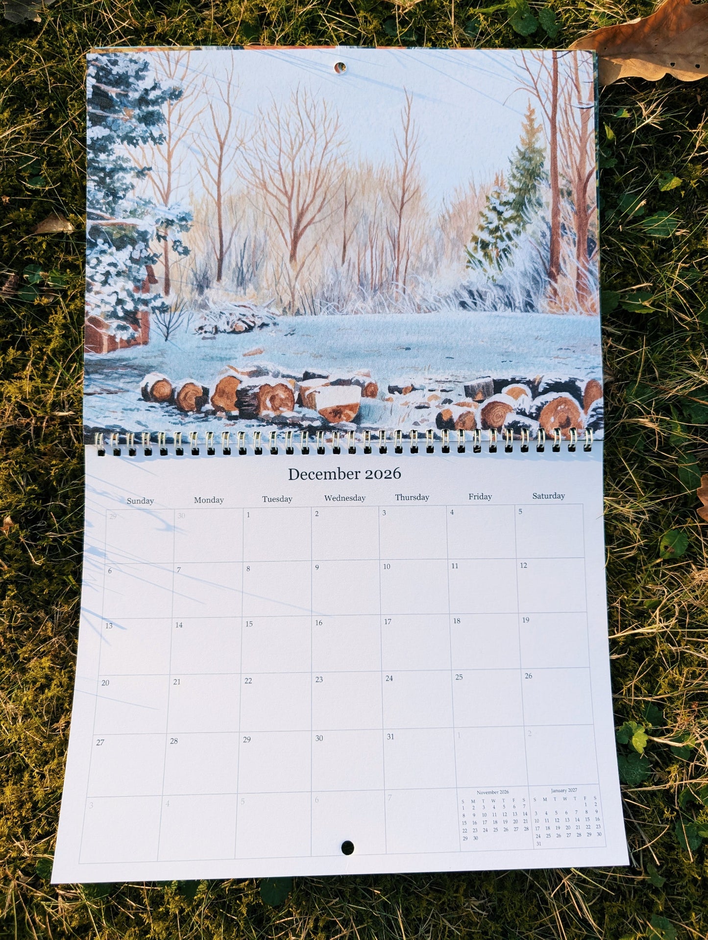 Calendar for December 2026 showing a winter landscape with trees and logs on grass.