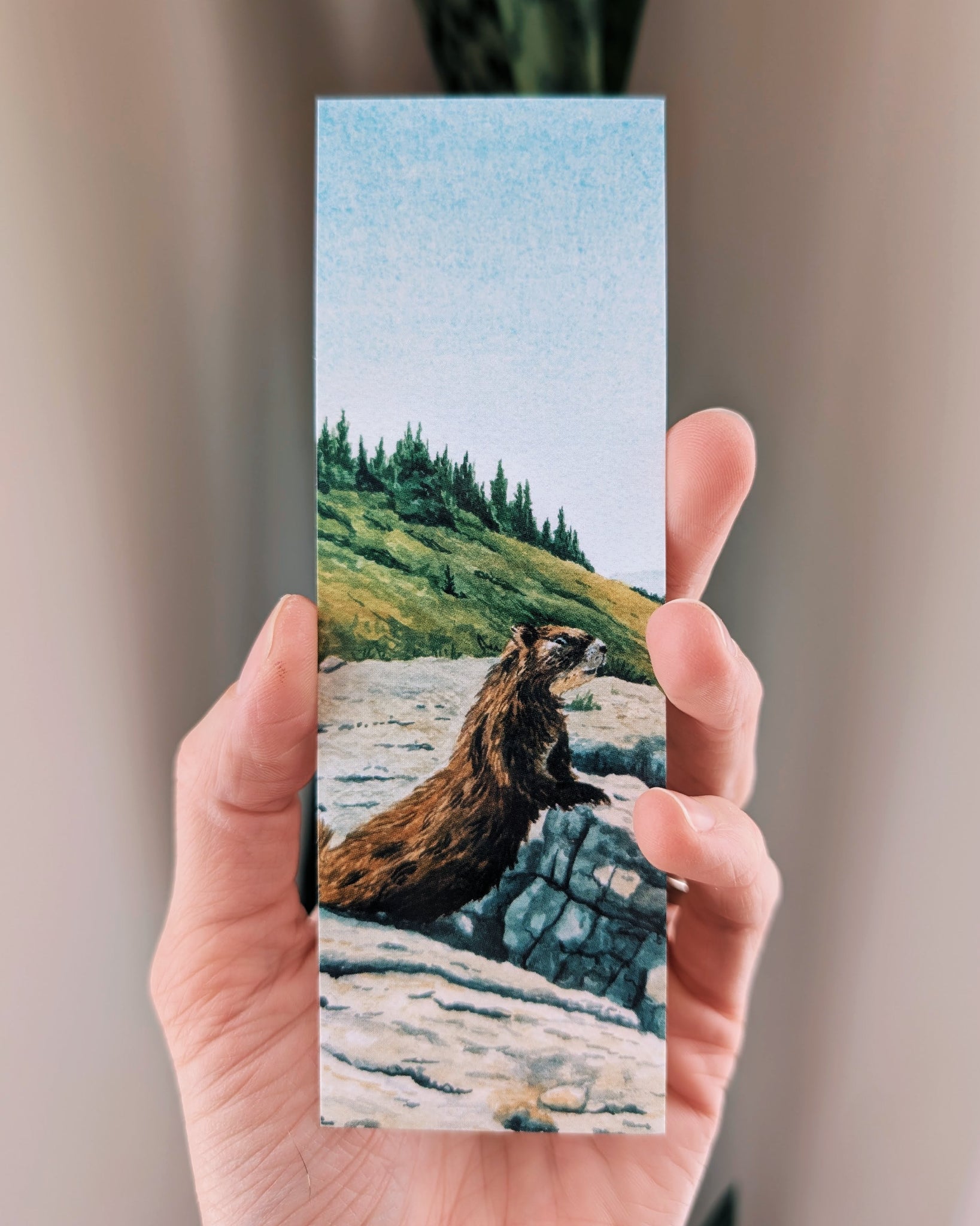 Hand holding a bookmark with a painted design of a marmot on a rock in a natural setting.