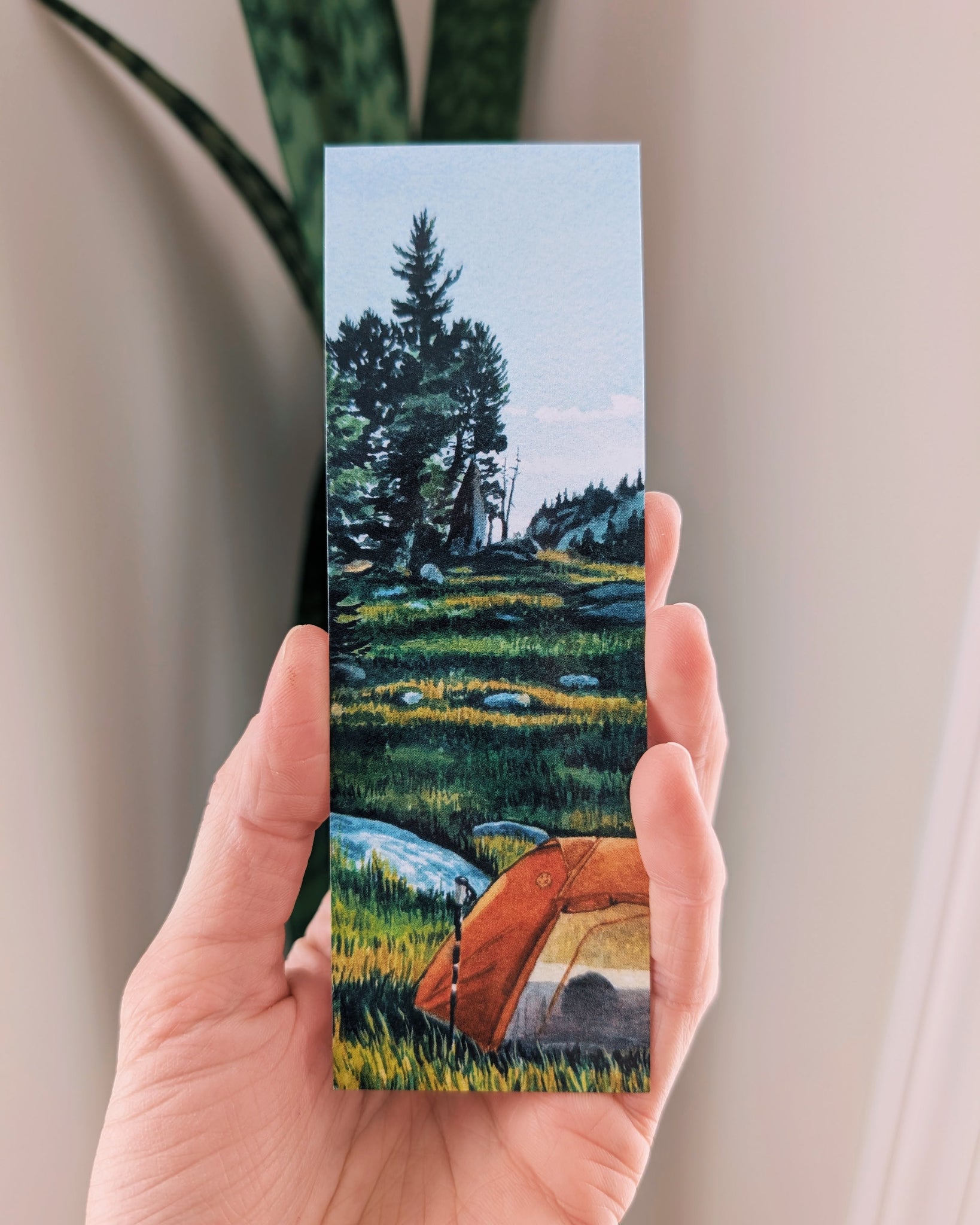 Hand holding a bookmark with an orange tent and a landscape design