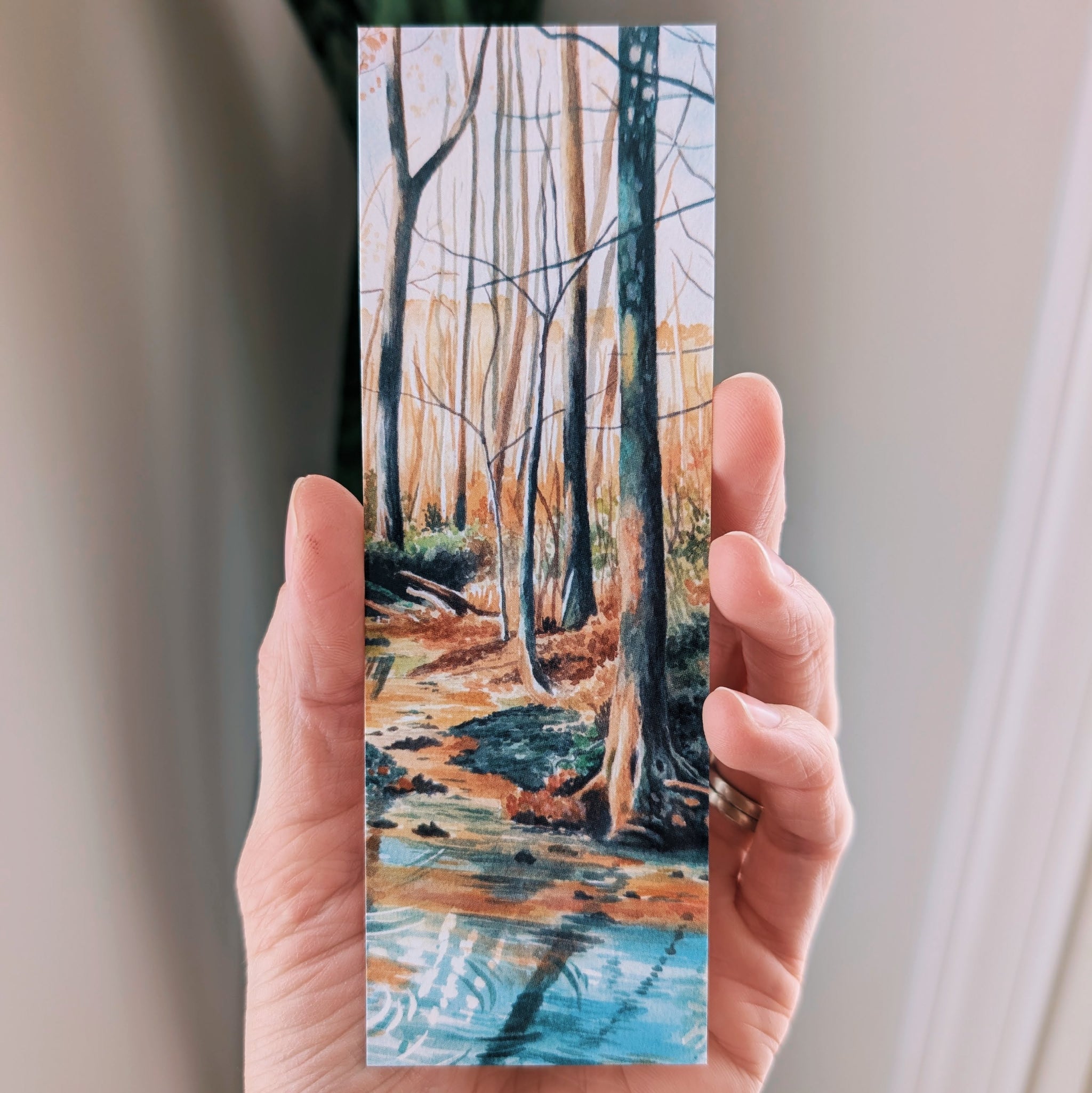 Hand holding a bookmark of a forest scene with trees and a stream