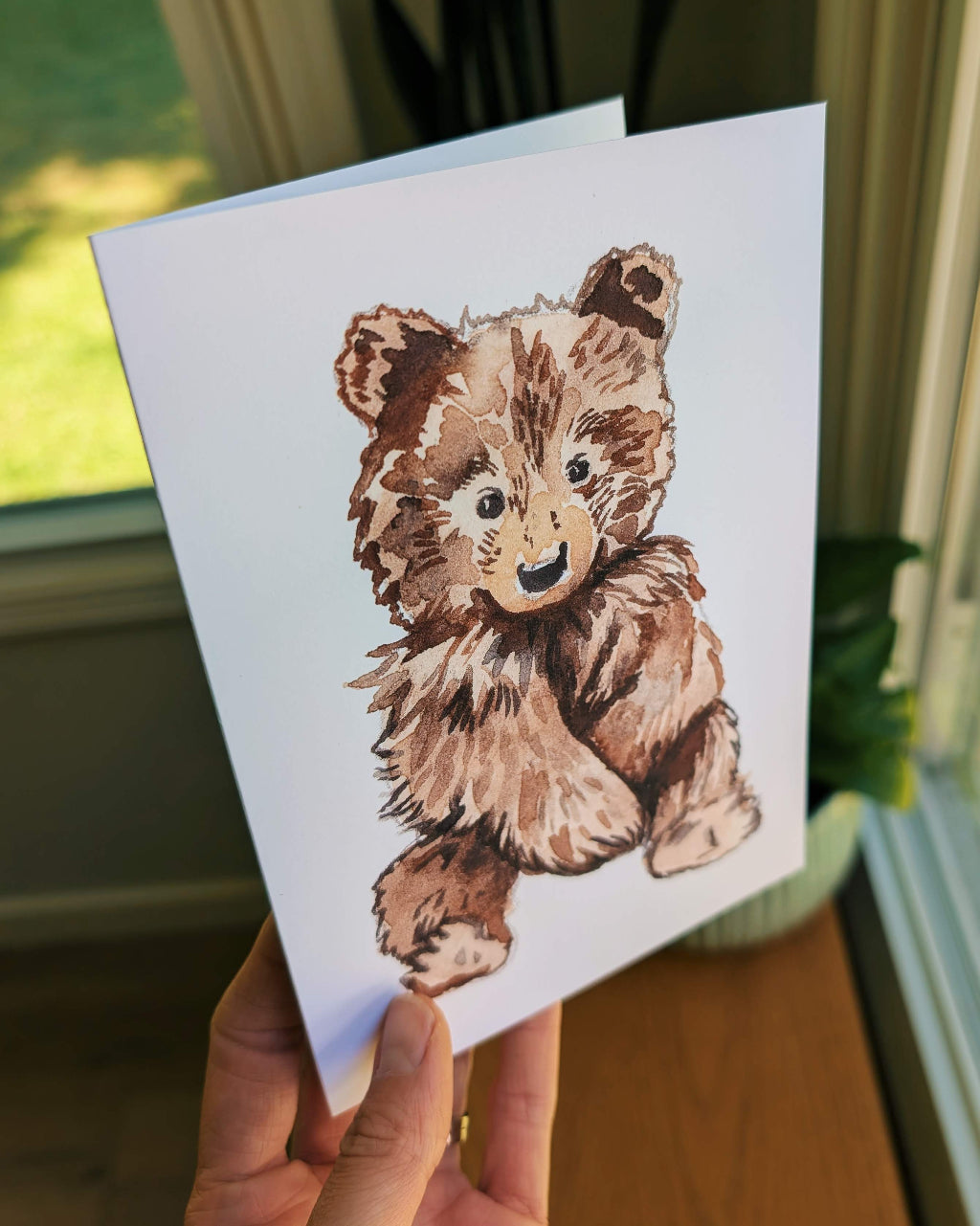 Card with a teddy bear illustration held by a hand indoors.