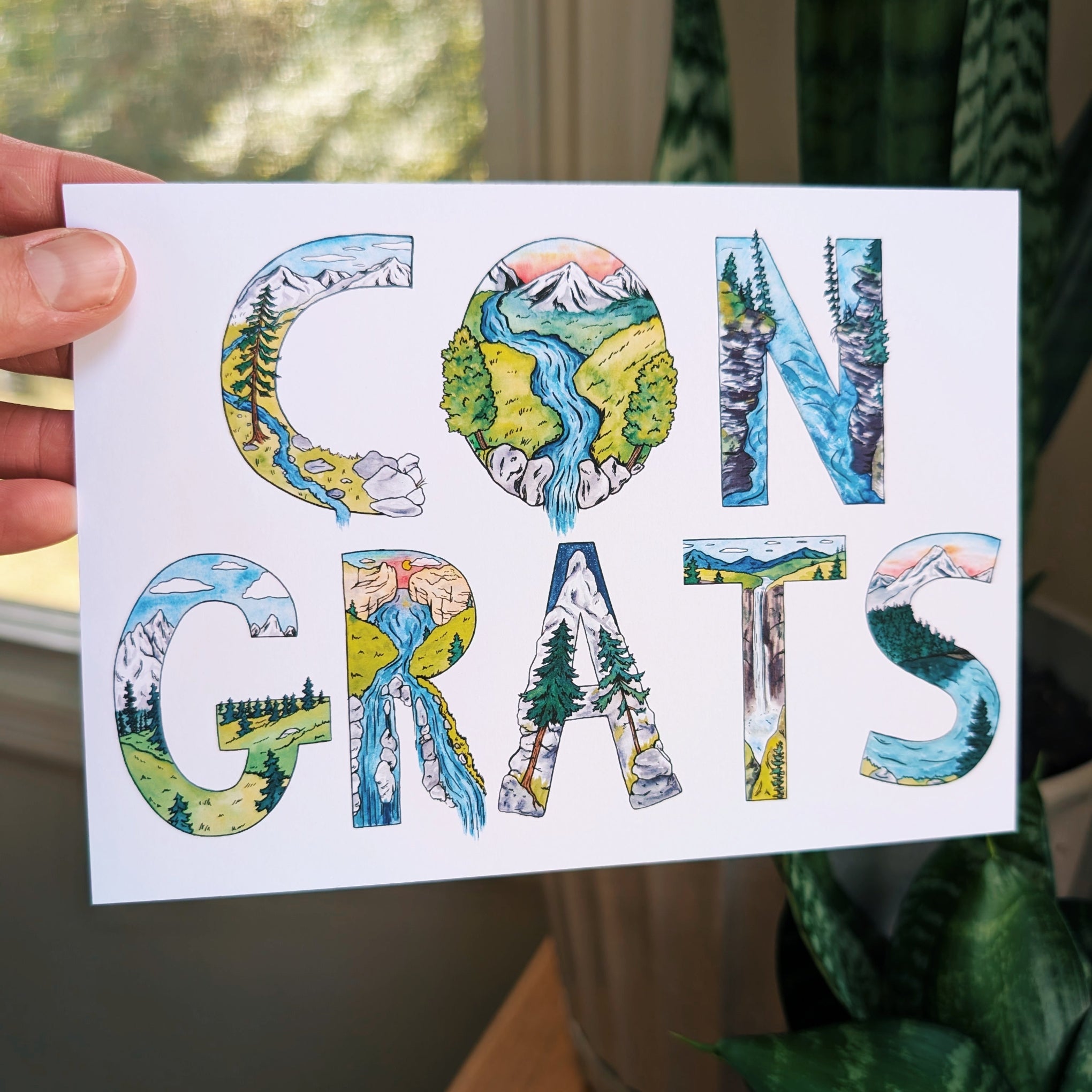 A hand holds a white greeting card with the word 'CONGRATS' spelled out in large illustrated letters filled with scenic landscape artwork. The background shows a softly blurred indoor setting with a window, curtain, and potted plant.