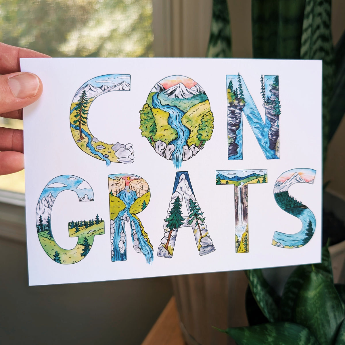 A hand holds a white greeting card with the word 'CONGRATS' spelled out in large illustrated letters filled with scenic landscape artwork. The background shows a softly blurred indoor setting with a window, curtain, and potted plant.