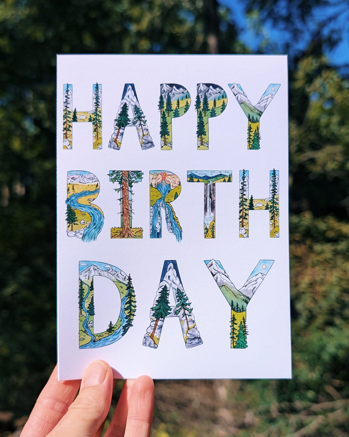 A hand holds a white greeting card that reads 'HAPPY BIRTH DAY' in large letters filled with scenic nature illustrations, set against a blurred outdoor background of trees and blue sky.