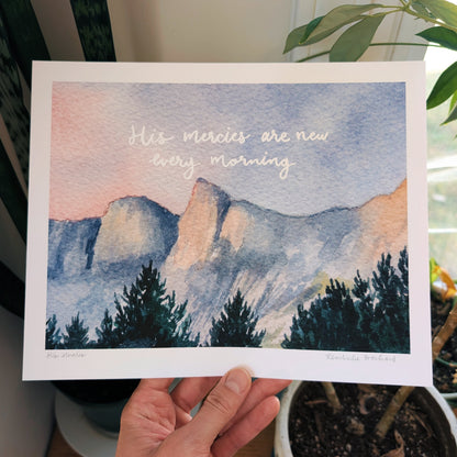 Hand holding a card with a mountain landscape and text 'His mercies are new every morning' against a plant background.