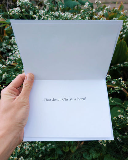 A hand holds an open greeting card outdoors. The card reads, 'That Jesus Christ is born!' in black serif text.