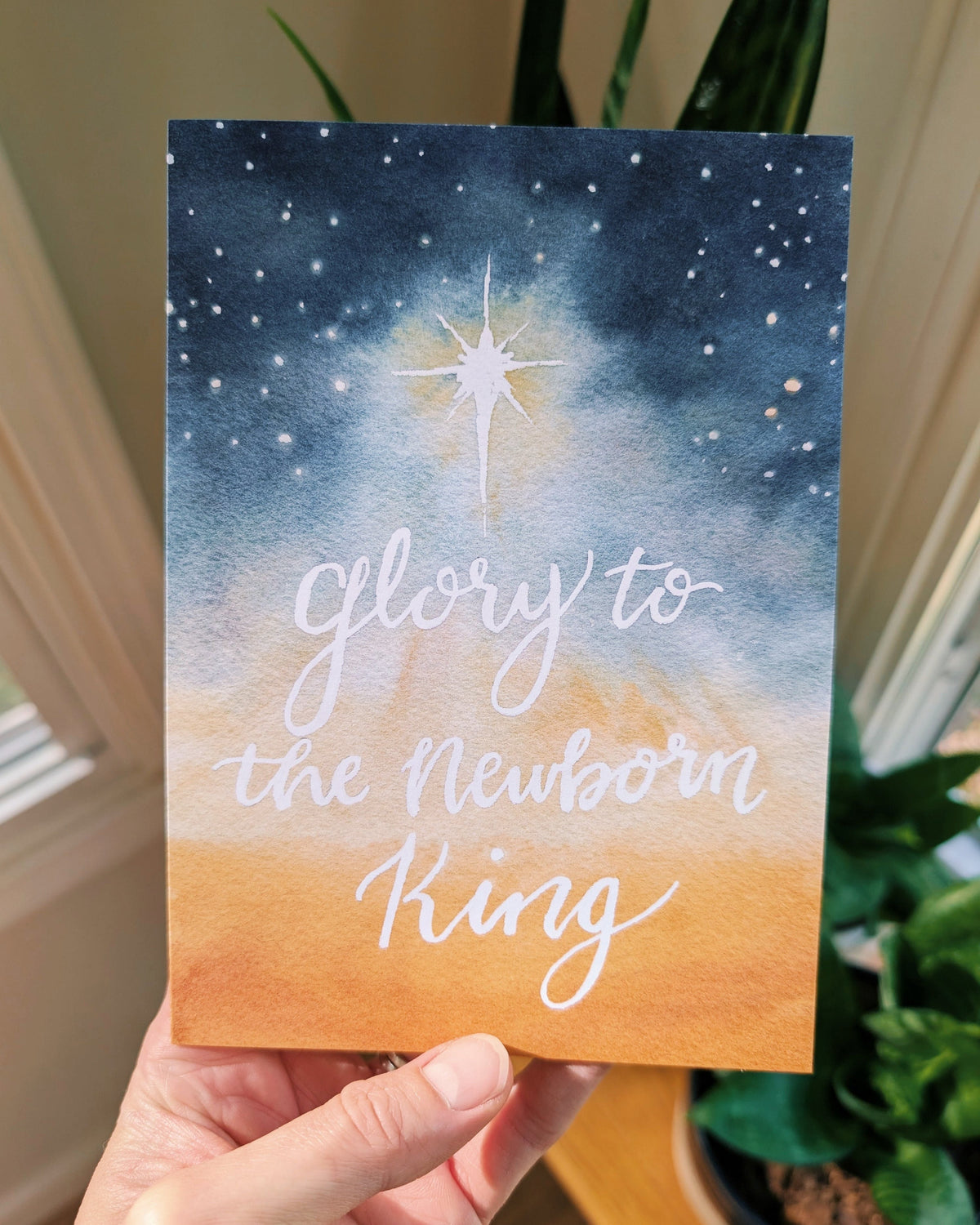A hand holds a card featuring a watercolor night sky with a bright star and the phrase 'Glory to the Newborn King' in white script.