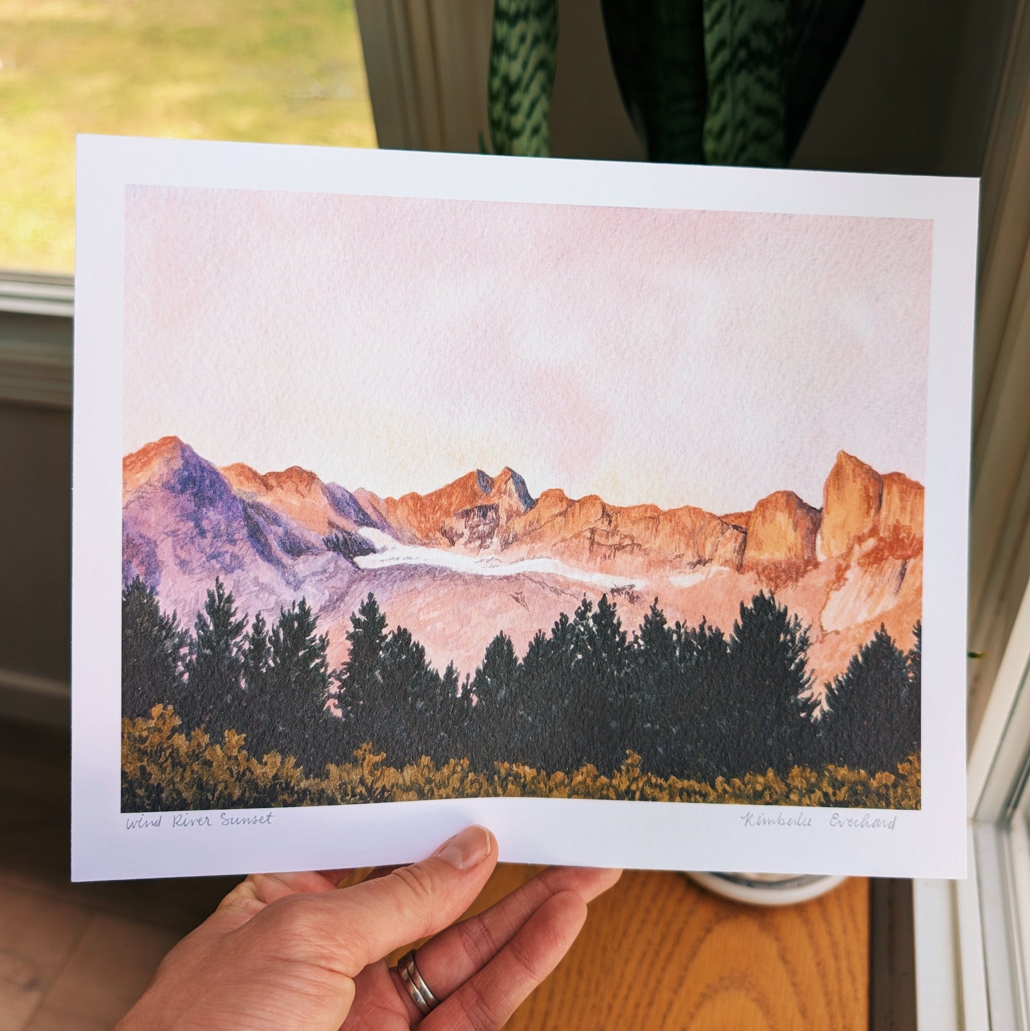 A hand holds a printed watercolor landscape artwork showing orange-brown mountains, a pale pink sky, dark evergreen trees, and yellow wildflowers.