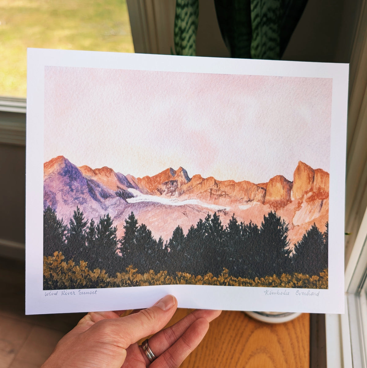A hand holds a printed watercolor landscape artwork showing orange-brown mountains, a pale pink sky, dark evergreen trees, and yellow wildflowers.