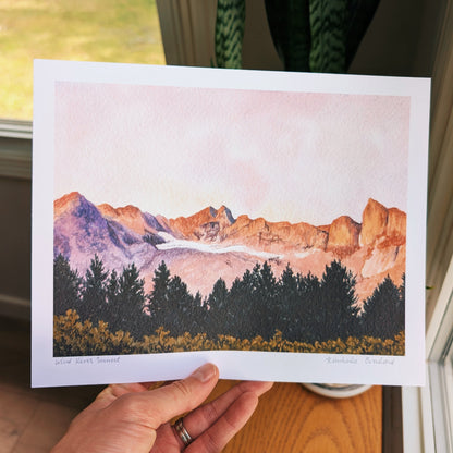 A hand holds a printed watercolor landscape artwork showing orange-brown mountains, a pale pink sky, dark evergreen trees, and yellow wildflowers.