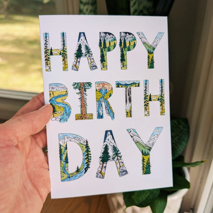 A hand holds a white greeting card with the words 'HAPPY BIRTH DAY' in large letters filled with a nature landscape pattern of trees, mountains, and water. The background shows a window, a wooden table, and a potted plant.