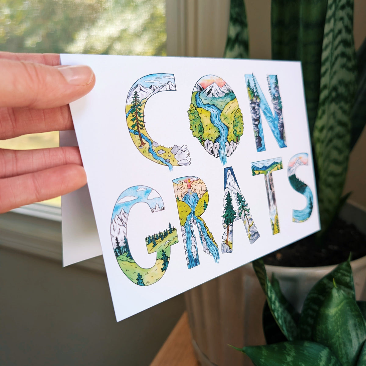 A hand holds a white greeting card that says 'CONGRATS' in large letters filled with colorful landscape illustrations, photographed indoors near a window and potted plants.